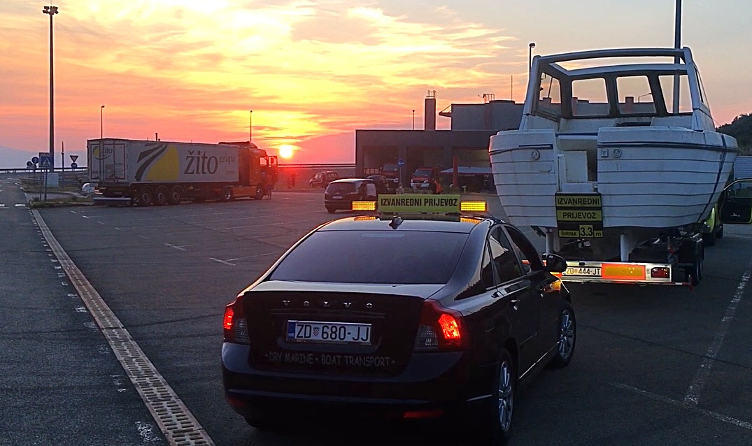 Black escort car with oversize transport sign driving behind a wide boat hull on a trailer at a Croatian highway rest area at sunset