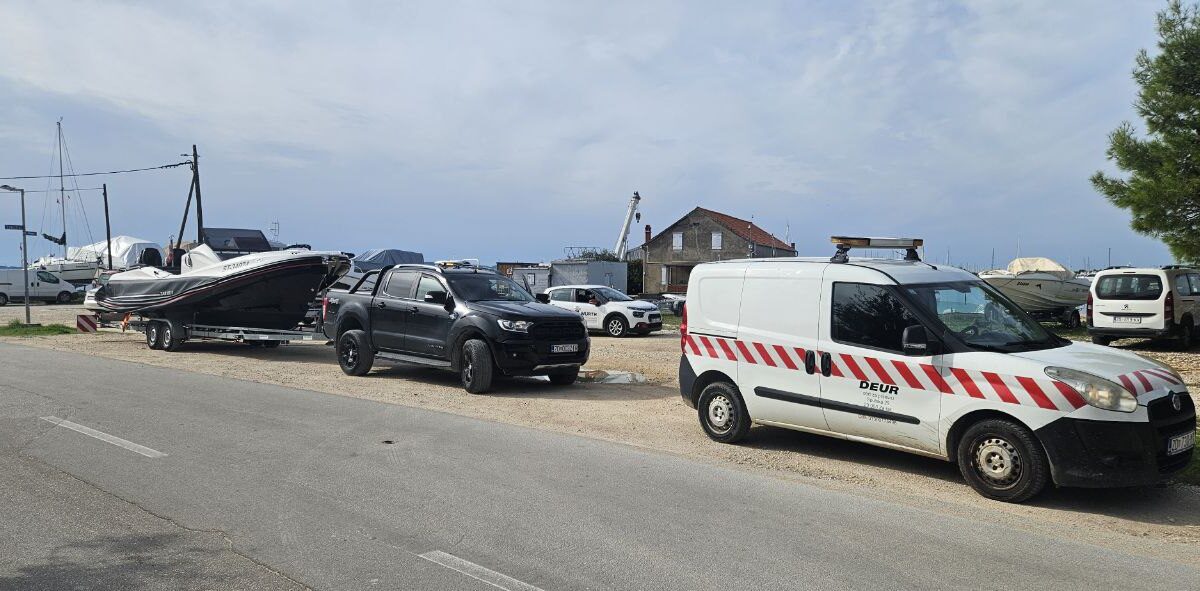 Black Ford Ranger towing a motorboat on a trailer with a white escort van parked beside the marina road
