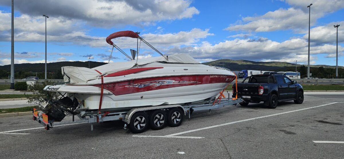 Black Ford Ranger towing a large red and white speedboat on a triple-axle trailer at a highway rest stop